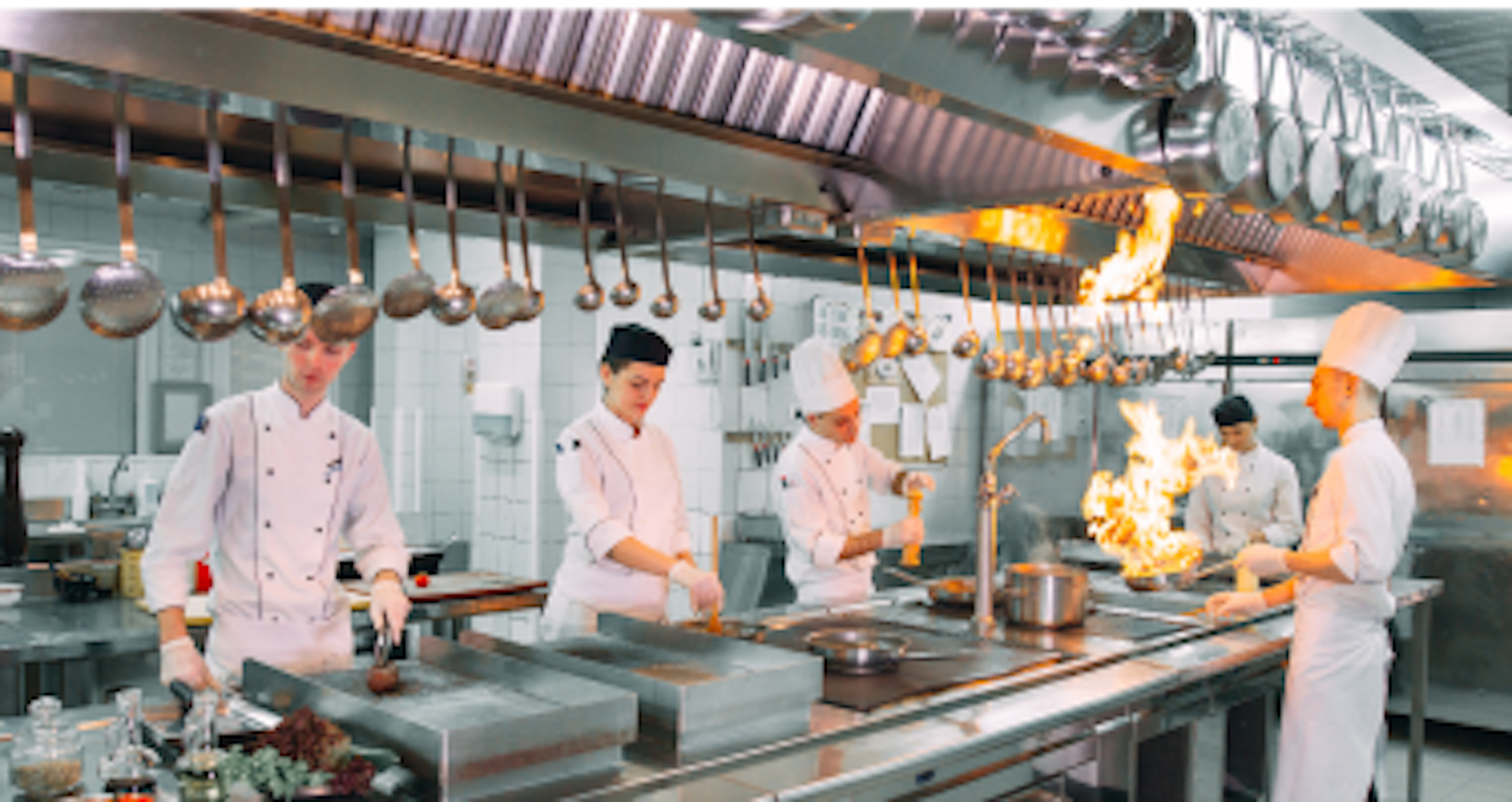 Four chefs in a professional kitchen, with one chef flambéing a dish on the stove.