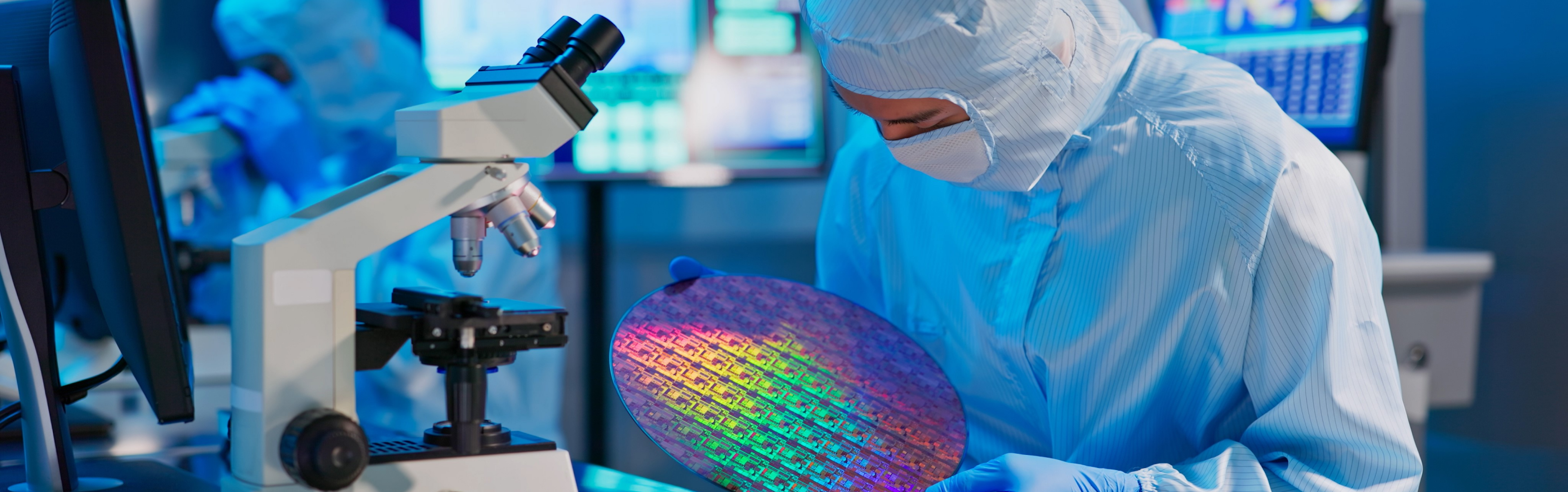 A person in a cleanroom suit examining a colorful silicon wafer under a microscope in a high-tech laboratory environment, indicating semiconductor manufacturing or research.