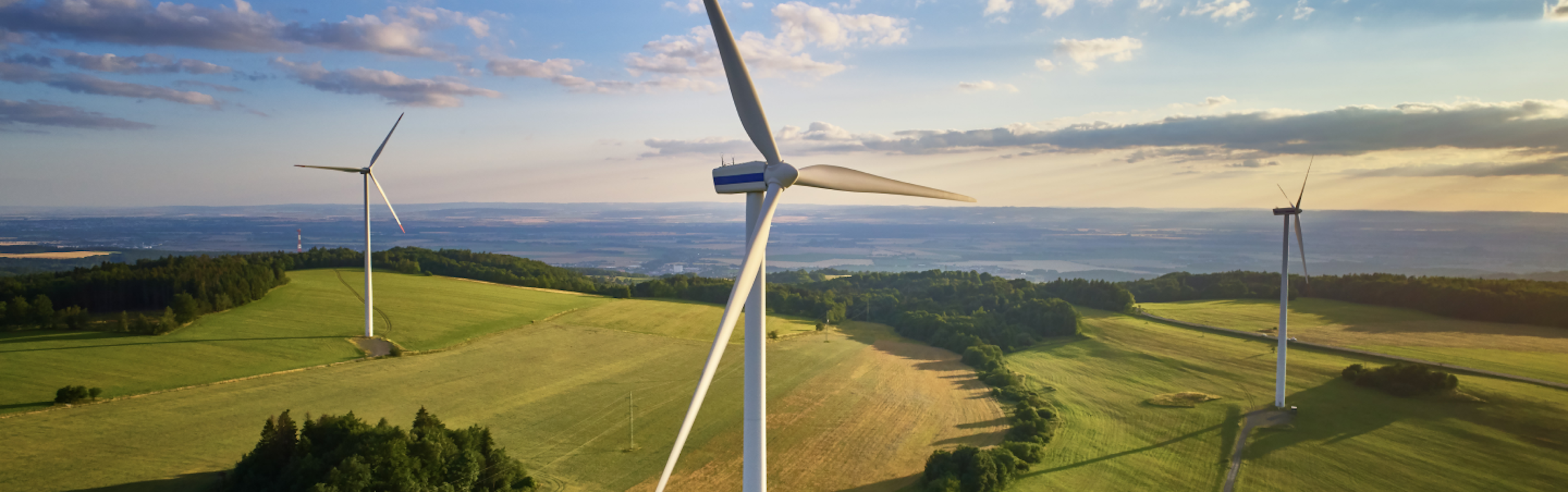 A scenic landscape with three large wind turbines on rolling green hills under a partly cloudy sky.