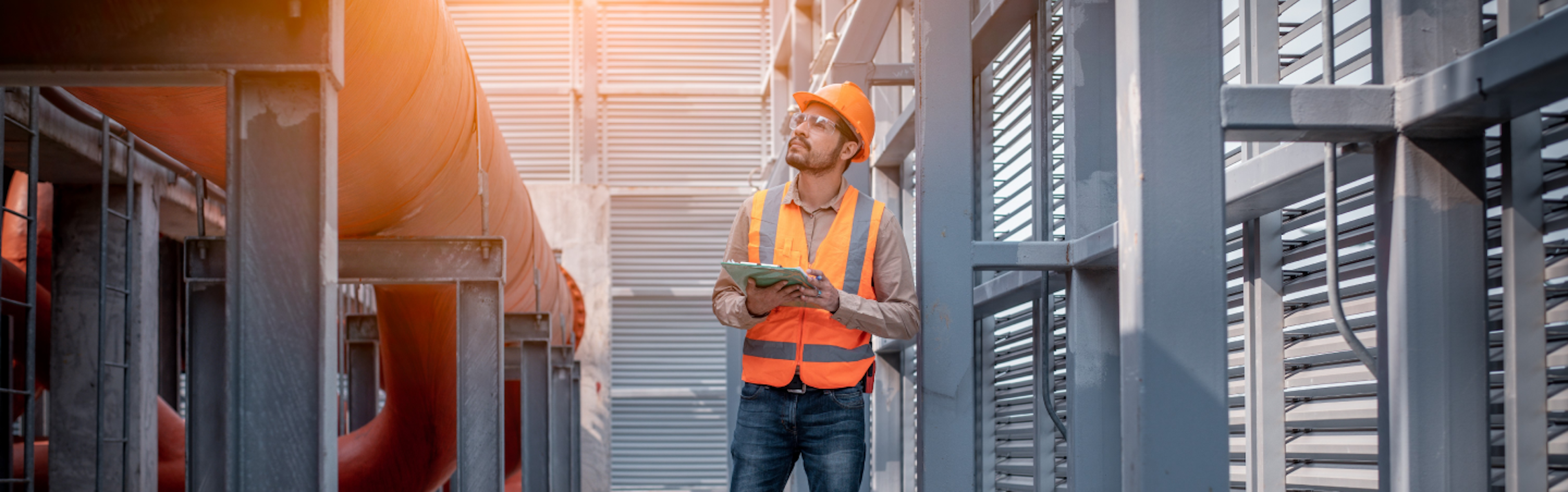 A construction worker wearing a hard hat, safety vest, and gloves is holding a tablet while standing on a metal staircase at a construction site.