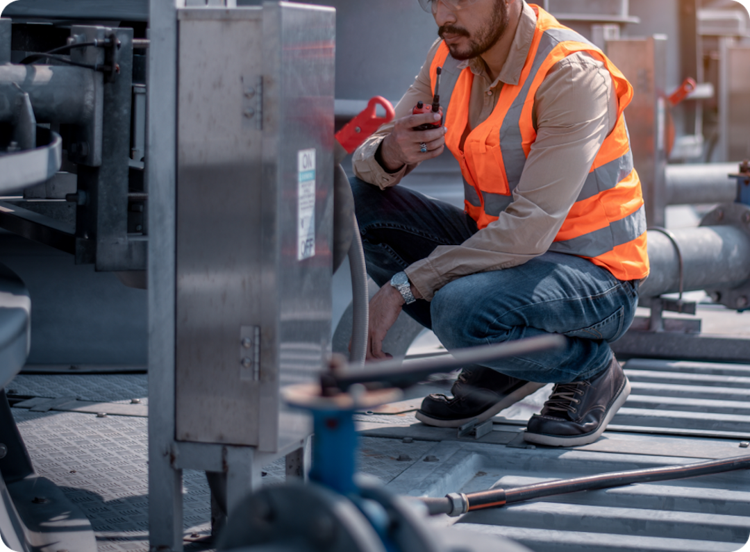 A worker in a reflective orange vest and safety gear is crouching while inspecting industrial equipment.