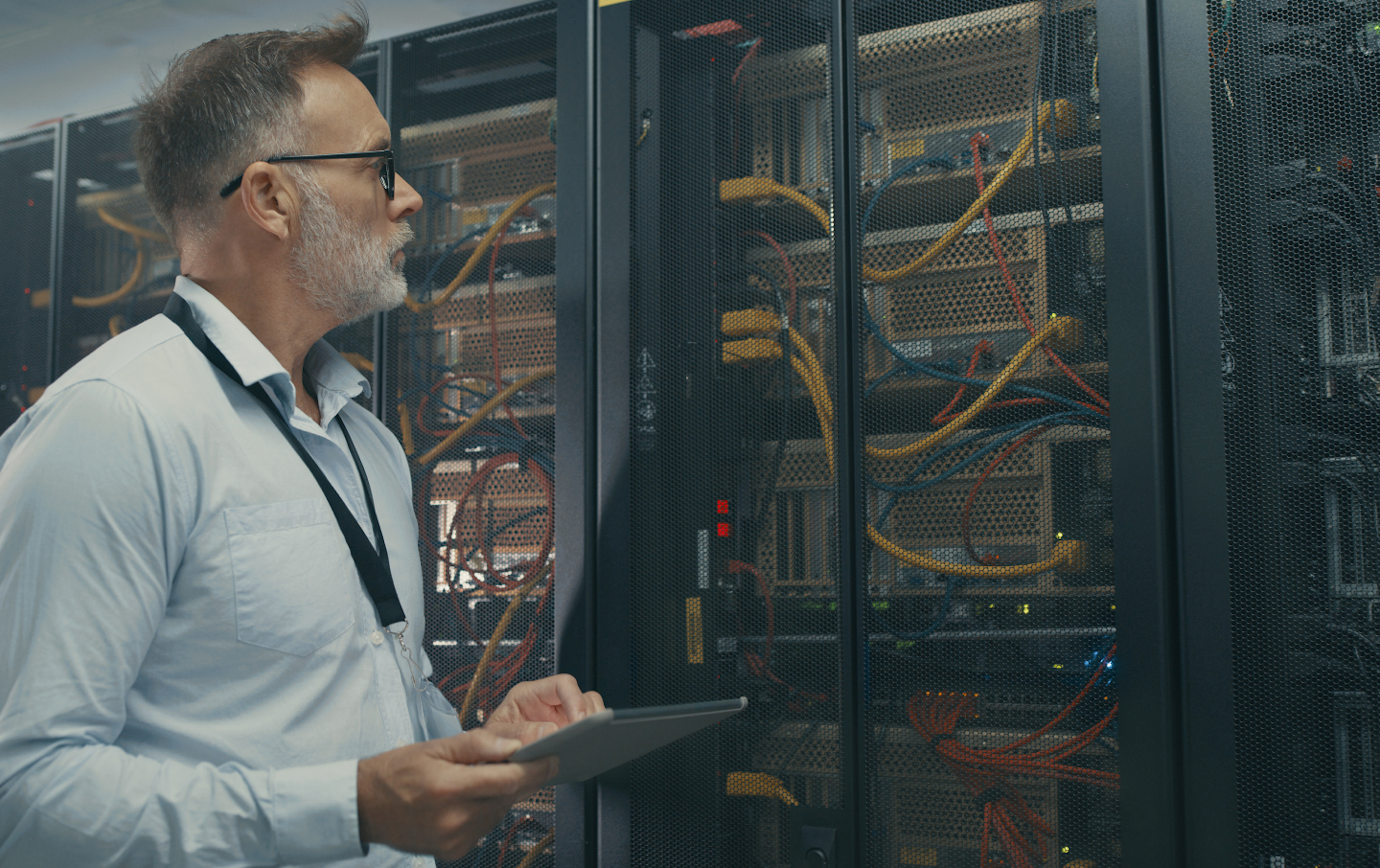 A man with glasses and a beard examines server racks in a data center, holding a tablet and wearing a lanyard, surrounded by networking cables and equipment.