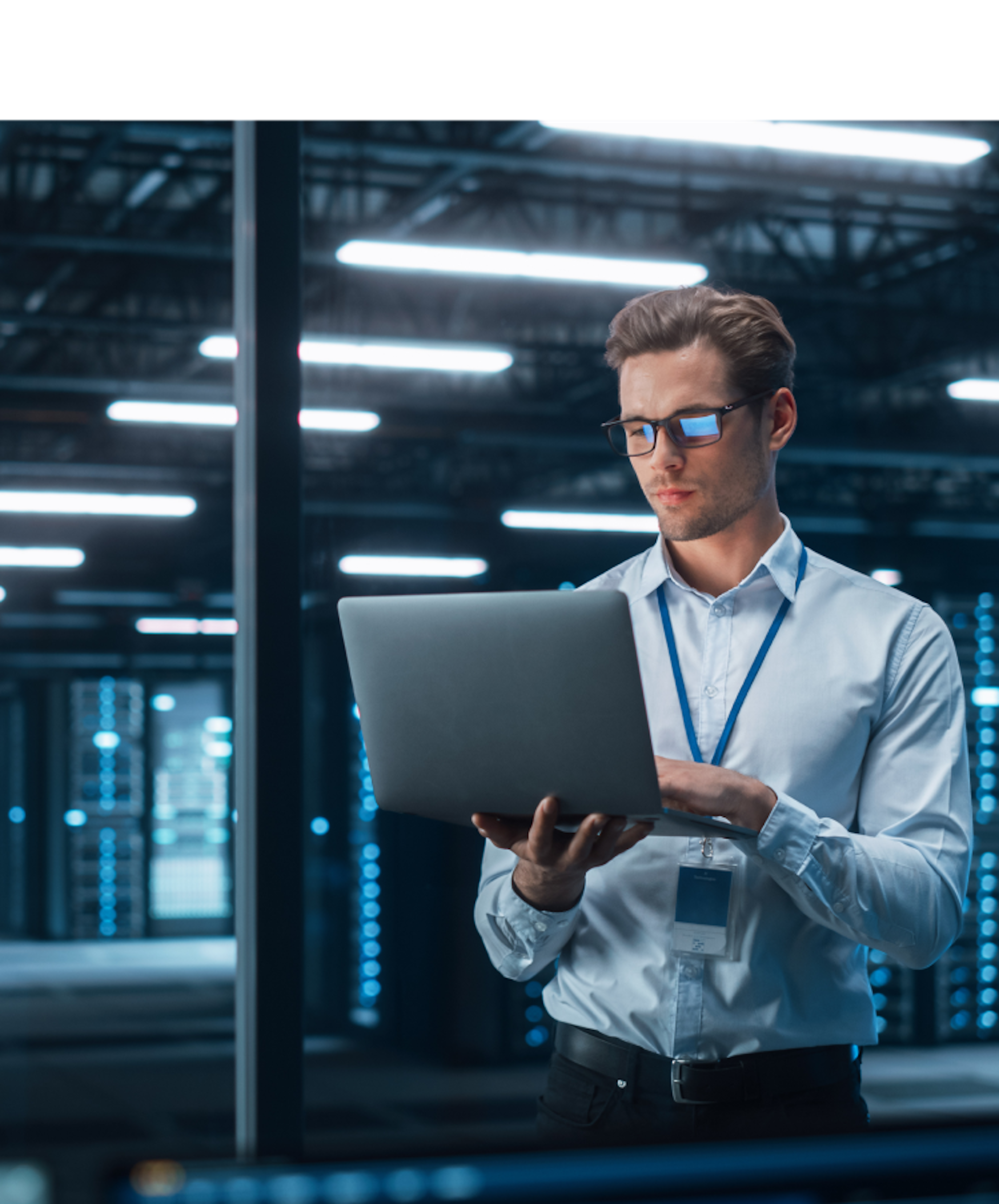 A man in a white shirt and glasses stands in a dimly lit server room, working on a laptop. He wears a lanyard and looks concentrated. Rows of server racks with blue lights are visible behind him.