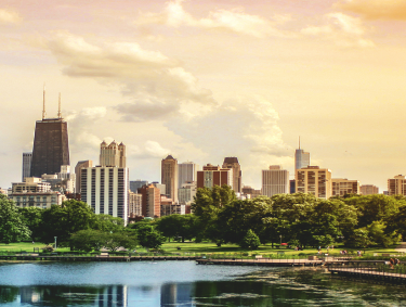 A city skyline with tall buildings is seen from across a green park under a cloudy sky.