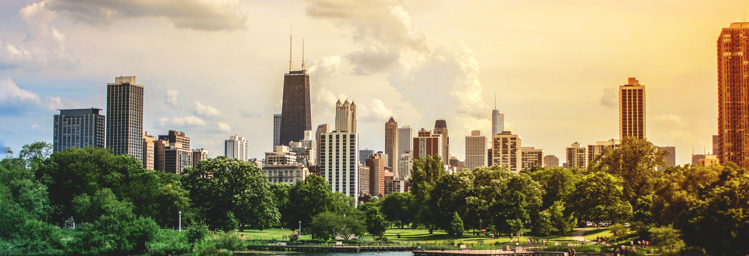 A city skyline with tall buildings is seen from across a green park under a cloudy sky.